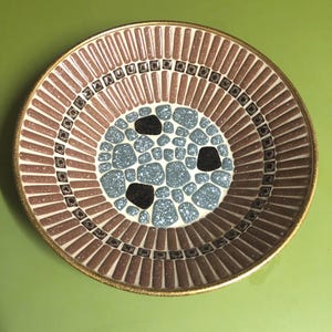 Könnte beinhalten: Eine runde Keramikschale mit einem zentralen Mosaikmuster aus hellblau und schwarz gesprenkelten Steinen. Der Rand besteht aus einem Ring kleiner dunkler Quadrate, gefolgt von einem breiten Band vertikaler, hellbrauner, gesprenkelter Fliesen.