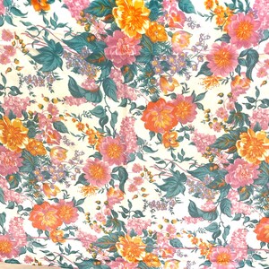 以下が含まれることがあります： クリーム色の背景に、オレンジ、ピンク、紫の花とティールグリーンの葉が密集したパターンで描かれた花柄の生地。さまざまな種類の花が繰り返されるデザインです。
