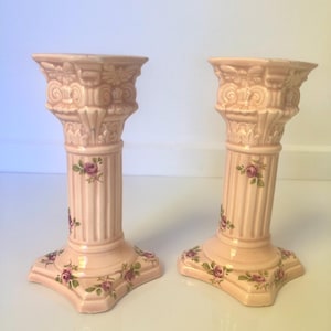 Op de afbeelding: Twee roze keramische kandelaars met een klassiek kolomontwerp. Elke houder heeft bloemaccenten met roze rozen en groene bladeren. De kandelaars hebben een vierkante basis en zijn ongeveer 20 cm hoog.