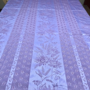 Pode incluir: Uma toalha de mesa lavanda com um padrão floral e de trigo. O design apresenta girassóis e outros elementos botânicos dispostos em faixas verticais. O tecido tem um brilho subtil, adicionando à sua aparência elegante.