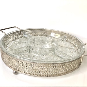 Peut inclure: Un plateau de service rond en métal argenté avec un motif latéral en maille décorative et deux poignées latérales. Le plateau comprend un insert en verre transparent amovible divisé en cinq sections avec un motif en étoile sur la base.