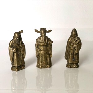 Peut inclure: Trois figurines antiques en laiton représentant des personnages asiatiques. Chaque figurine est détaillée avec des vêtements et des traits du visage traditionnels. Les figurines sont posées sur une surface blanche. Les figurines sont dans diverses poses, dont une tenant une coupe.
