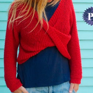 Könnte beinhalten: Ein roter Strickpullover mit einem Kreuz-Detail auf der Vorderseite. Der Pullover ist kurz und hat lange Ärmel. Das Model trägt den Pullover über einem blauen Shirt und Jeans. Der Text "uKnitMe2" ist auf der linken Seite des Bildes sichtbar.