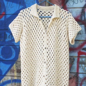 May include: A cream-coloured, short-sleeved crochet shirt with a collared neckline and button-down front. The shirt is hanging on a blue hanger against a graffiti-covered wall. The text "UknitMe2" is at the top.