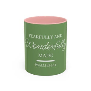 May include: A ceramic coffee mug with a green exterior and a pink interior. The mug features the text "FEARFULLY AND Wonderfully MADE" in white script, along with "PSALM 139:14". The mug is cylindrical and suitable for hot or cold beverages.