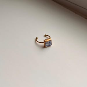 May include: Gold-toned adjustable ring featuring a square, blue and white patterned ceramic tile. The ring has a delicate, open design with small ball accents on the ends.