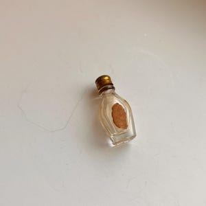 May include: A miniature clear glass bottle with a rounded body and a narrow neck. The bottle is topped with a small, ribbed metallic gold screw cap and features a single, plain, light brown oval-shaped paper label centred on the front surface.