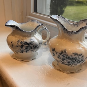 May include: Two cream-colored ceramic pitchers featuring a blue floral pattern and a blue speckled rim. Each pitcher has a scalloped base and a curved, flared spout. The pitchers are glazed with a smooth finish and have a rounded, bulbous body shape.