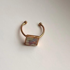 May include: A gold-colored ring with a rectangular floral design. The ring has a curved band with small ball accents on each end. The floral design features red, blue, and yellow hues.