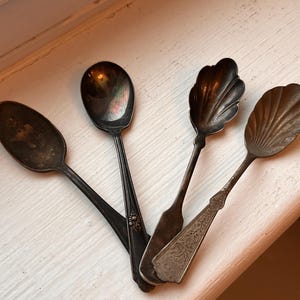 May include: A set of four tarnished silver-toned metal spoons. Two spoons feature oval bowls, while the other two have shell-shaped bowls with ridged detailing. The handles exhibit varied designs, including floral engravings and smooth, tapered shapes.
