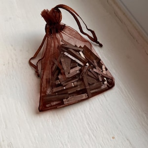 May include: A small, brown, semi-sheer organza drawstring pouch filled with numerous thin, rectangular, metallic-toned pieces. The bag is gathered at the top with matching brown ribbon ties, containing a collection of small, flat, rigid components.