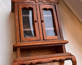 May include: A miniature wooden cabinet featuring two glass-paneled doors with small knobs, an open shelf below the doors, and four curved legs. The piece is finished in a medium brown wood tone with decorative carved detailing at the top.