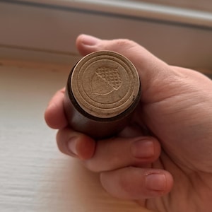 May include: A cylindrical wooden stamp featuring a circular face with an engraved acorn design. The stamp has a dark brown wooden handle and a light tan circular base with a detailed, etched acorn icon centered on the flat surface.