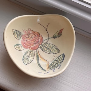 Könnte beinhalten: Eine cremefarbene Keramikschale mit unregelmäßiger, abgerundeter Form. Das Innere zeigt eine handgezeichnete Illustration einer rosa Rose mit grünen, schraffierten Blättern und einer kleinen Knospe im skizzenhaften Stil mit sichtbaren Strichen.