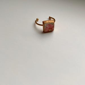 May include: A gold-toned adjustable ring featuring a square floral design. The ring's band is open, with small ball accents on each end. The square centerpiece displays a pink and white floral pattern.