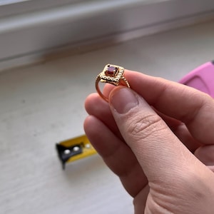 May include: A gold ring featuring a square-shaped setting with a central deep red gemstone surrounded by a textured border. The ring is held near a yellow measuring tape, though the specific measurements are not clearly visible in the image.