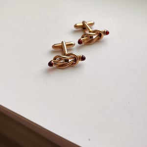 May include: A pair of gold-toned cufflinks featuring a central knot design. Each end of the knot is accented with a small, round, dark red stone. The cufflinks include a standard T-bar swivel mechanism for securing them through shirt cuffs.