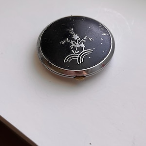 May include: A round, black compact mirror with a silver-toned metal rim and a small gold-colored latch. The top surface features a delicate, engraved silver floral design with a central flower and curved lines, showing signs of wear.