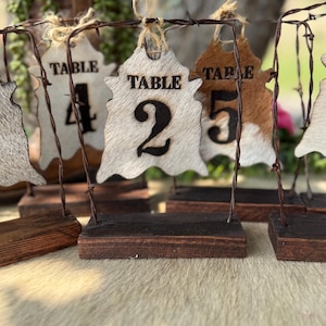 May include: A set of table number signs featuring hide-shaped cutouts with hair-on-hide texture in white, brown, and tan. Each sign displays the word TABLE and a large black number, suspended by twine from a dark brown barbed wire metal frame.
