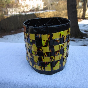 May include: A black and yellow woven basket with a flame pattern. The basket is made of duct tape and is sitting on a white surface.