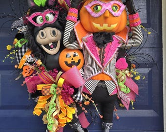 May include: A Halloween wreath featuring a pumpkin-headed figure in a striped suit and top hat, alongside a black donkey head wearing a pink mask. The arrangement includes orange, pink, and black ribbons, small pumpkins, and floral accents.