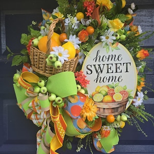 May include: A vibrant, decorative wreath featuring a basket overflowing with oranges and lemons, accented with daisies and greenery. A large green bow and a sign that reads "home SWEET home" complete the design. The wreath is a cheerful, welcoming home decor item.