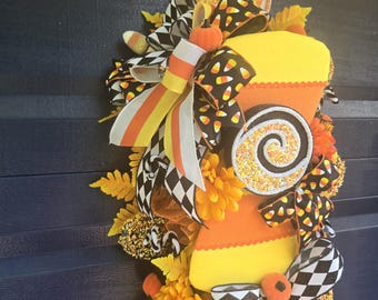 May include: A festive Halloween wreath featuring a large candy corn design. The wreath is adorned with black and white checkered bows, orange and yellow flowers, and candy corn accents. A large swirl candy and small pumpkins add to the holiday theme.