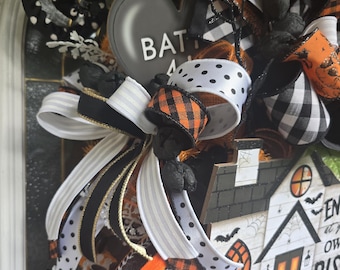 May include: A Halloween wreath featuring a house sign with text, black and white polka dot ribbons, orange and black plaid ribbons, and white striped ribbons. The wreath includes black glittery branches, white flowers, and a gray heart-shaped sign with the text BAT.
