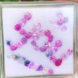 May include: A collection of loose, faceted gemstones in various shades of pink, purple, and blue, arranged in a clear plastic display box with a white foam insert. The stones feature diverse shapes including oval, round, square, and pear cuts.