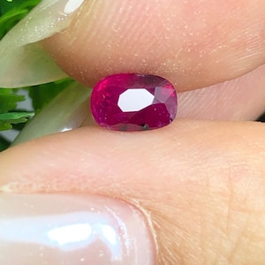 Op de afbeelding: Een robijn edelsteen in kussenslijpvorm met een diepe, verzadigde magenta-rode kleur en meerdere facetten die het licht reflecteren. De steen heeft een helder, gepolijst oppervlak met zichtbare interne insluitingen en een symmetrische vorm.