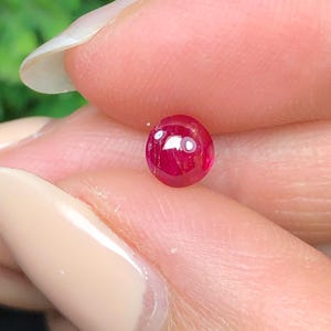 May include: A small, round, cabochon-cut gemstone with a vibrant, deep pinkish-red colour. The stone features a smooth, polished surface that reflects light, showcasing its translucent quality and rich, saturated hue in a circular shape.