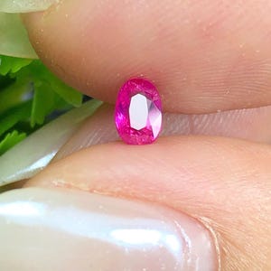 May include: A small, oval-cut gemstone with a vibrant, saturated hot pink color. The stone features multiple facets that reflect light, creating a brilliant, sparkling appearance. It is held between two fingers, showcasing its translucent quality.