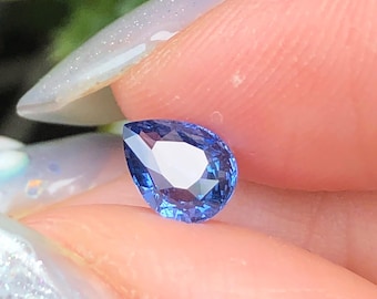 Puede incluir: Una piedra preciosa de zafiro azul en forma de pera con una superficie facetada que refleja la luz. La piedra muestra un color azul profundo y vibrante y una calidad transparente, exhibiendo cortes geométricos precisos.