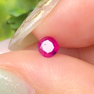 May include: A small, round, faceted gemstone with a vibrant, deep pinkish-red color. The stone features a brilliant cut with multiple triangular facets that reflect light, creating a clear, sparkling appearance on its surface.