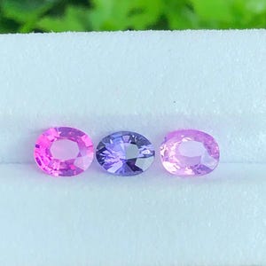 Könnte beinhalten: Drei oval geschliffene Edelsteine in einer horizontalen Reihe. Der linke Stein ist leuchtend pink, der mittlere Stein ist tief violettblau und der rechte Stein ist zartrosa. Jeder Edelstein hat eine facettierte Oberfläche, die das Licht reflektiert.