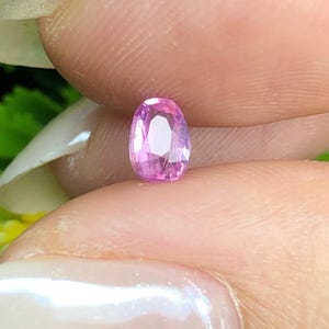 Puede incluir: Una pequeña piedra preciosa de color rosa con forma ovalada y superficie facetada. La piedra muestra un tono rosa vibrante y translúcido, tallada con múltiples facetas geométricas que reflejan la luz, creando un aspecto brillante.