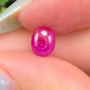 Puede incluir: Una pequeña piedra preciosa ovalada de color rosa intenso vibrante con una superficie de cabujón lisa y pulida. La piedra muestra una calidad translúcida con reflejos de luz visibles en su parte superior redondeada, exhibiendo un color magenta.