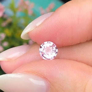 May include: A round, light pink gemstone with multiple facets, held between fingers. The gem has a brilliant cut, reflecting light. The background is blurred, with hints of pink and green. The gemstone is a delicate, feminine piece.