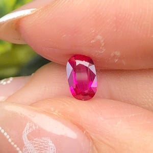 Op de afbeelding: Een levendige, ovaal geslepen robijn met een diepe, verzadigde roze-rode kleur en meerdere facetten die het licht reflecteren. De steen is klein en gepolijst, met een heldere, transparante kwaliteit en scherpe randen in een symmetrische vorm.
