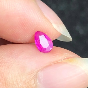 May include: A small, pear-shaped, faceted gemstone with a vibrant, saturated hot pink color. The stone features a polished surface with multiple triangular and diamond-shaped facets that reflect light, creating a bright, clear appearance.