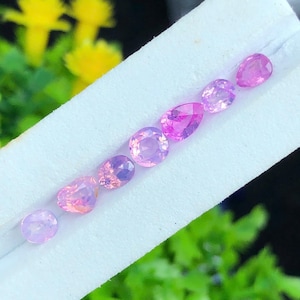 May include: A collection of seven faceted pink gemstones arranged in a diagonal line on a white display card. The stones vary in shape, including round, oval, and pear cuts, each exhibiting a translucent quality and vibrant pink color tones.