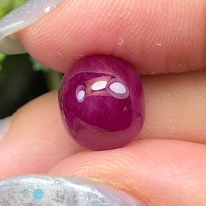 May include: A smooth, oval-shaped, deep magenta-coloured gemstone with a polished, reflective surface. The stone exhibits a rich, saturated hue and a rounded cabochon cut, catching light with visible white highlights on its curved top surface.
