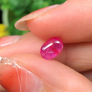 May include: A vibrant, oval-shaped, pink gemstone with a polished surface. The gem is held between fingers, showcasing its clarity and colour. The background is blurred, highlighting the gemstone's details.
