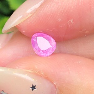May include: A pear-shaped, faceted pink gemstone held between fingers. The gem has a bright, light pink color and is cut to reflect light. The background is blurred, with hints of green foliage.