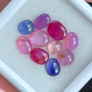 May include: A collection of ten oval-shaped, polished gemstones in a white display box. The stones feature a variety of colors including deep blue, vibrant magenta, soft pink, and light purple, with some showing internal natural inclusions.