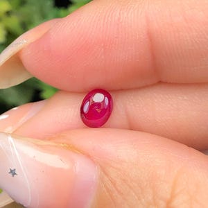 May include: A small, oval-shaped, deep red gemstone with a smooth, polished cabochon surface. The stone exhibits a vibrant, saturated crimson hue and a reflective, glossy finish that catches the light, highlighting its rounded, dome-like profile.