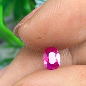 Puede incluir: Una gema de rubí facetada de corte ovalado. La gema es de un color rojo rosado vibrante, con múltiples facetas que reflejan la luz. El rubí se sostiene entre dos dedos, mostrando su superficie pulida y claridad.