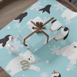 May include: A wrapped gift with a brown ribbon and bow sits on light blue wrapping paper. The paper features a pattern of penguins, polar bears, and snowflakes. Scissors and tape are also visible.