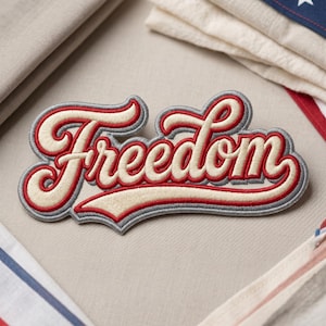 May include: Embroidered patch with the word "Freedom" in a stylized script. The lettering is cream-colored with red and gray accents, creating a vintage aesthetic. The patch is likely for clothing or accessories.