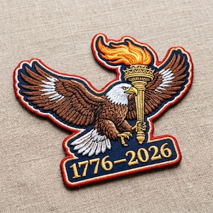 May include: Embroidered patch featuring a detailed eagle with outstretched wings, holding a golden torch with a flame. The patch has a blue background with the text "1776-2026" in gold. The eagle is brown, white, and gold, with a red border.